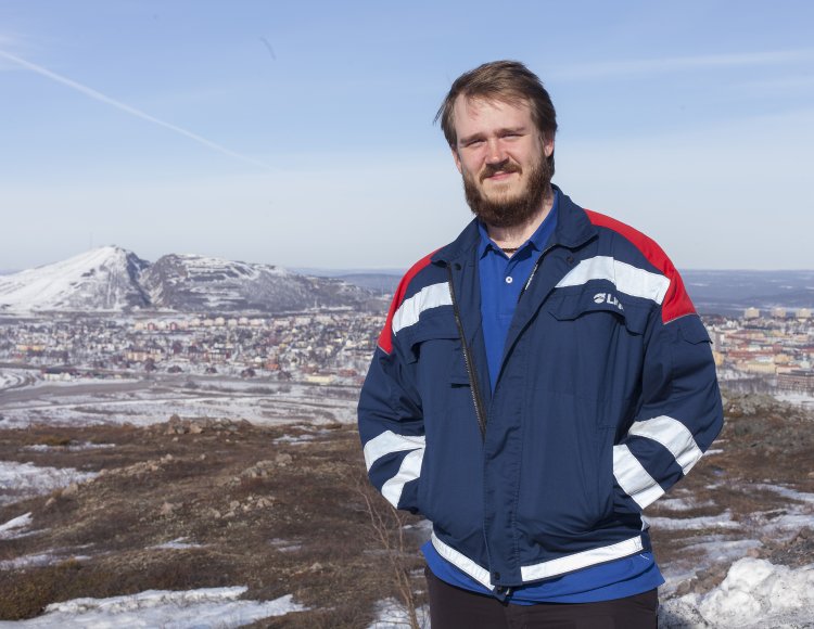 Petter Madsen jobbar som utvecklingsingenjör på LKAB. Han står högt upp på ett delvis snötäckt fjäll. Bakom honom skymtar man Kiruna. Solen skiner på en blå himmel. 