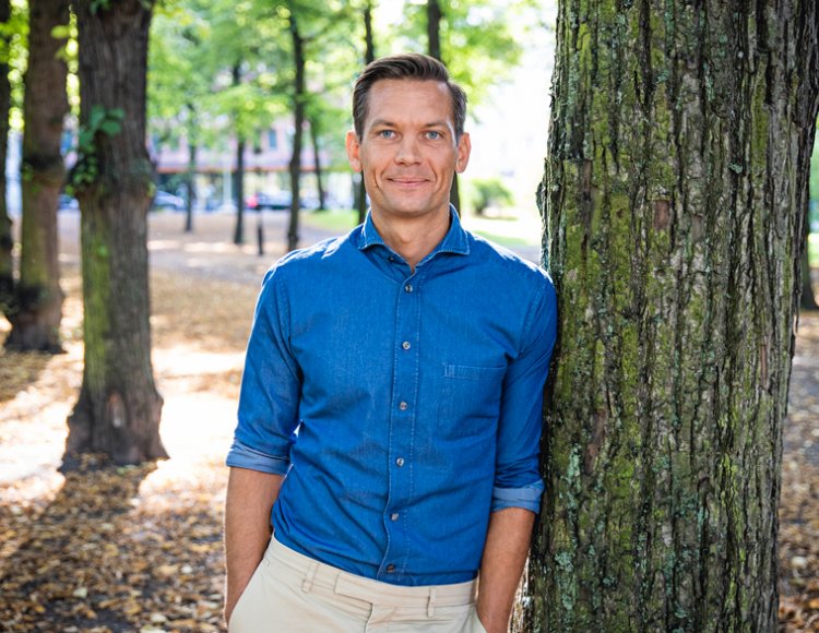 Rikard Nyhren står i blå skjorta utomhus och lutar sig mot ett träd. 