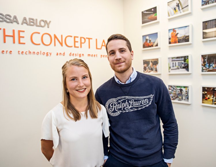 André More och Emelie Öhrn ståendes tillsammans på Assa abloys ljusa kontor. I bakgrunden sitter inramade bilder på väggarna samt företagets slogan. 