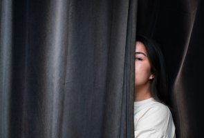 En bild med en tjej som står halvt gömd bakom en svart gardin.