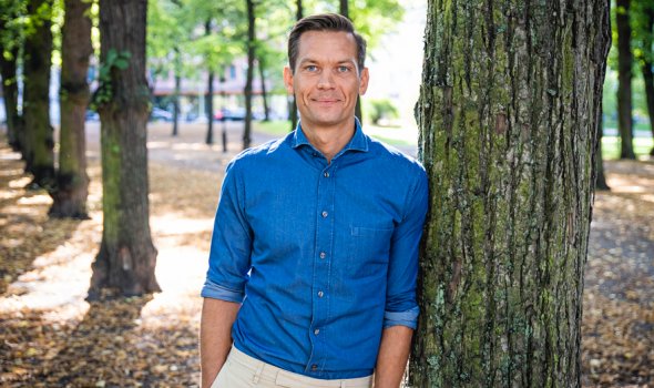 Rikard Nyhren står i blå skjorta utomhus och lutar sig mot ett träd. 