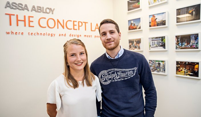 André More och Emelie Öhrn ståendes tillsammans på Assa abloys ljusa kontor. I bakgrunden sitter inramade bilder på väggarna samt företagets slogan. 