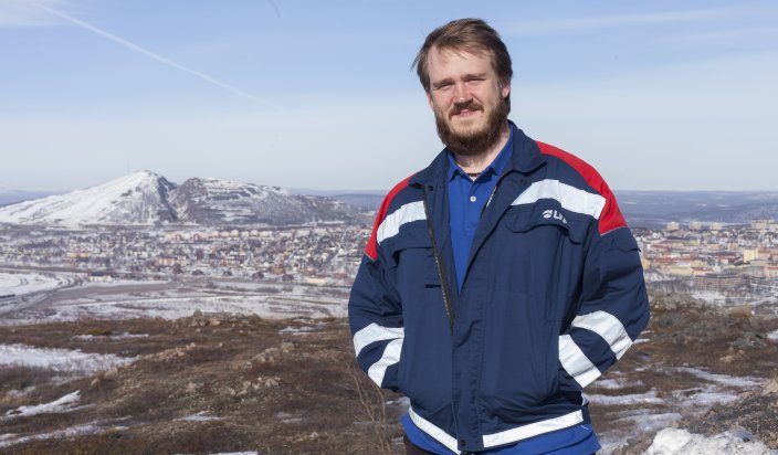 Petter Madsen jobbar som utvecklingsingenjör på LKAB. Han står högt upp på ett delvis snötäckt fjäll. Bakom honom skymtar man Kiruna. Solen skiner på en blå himmel. 