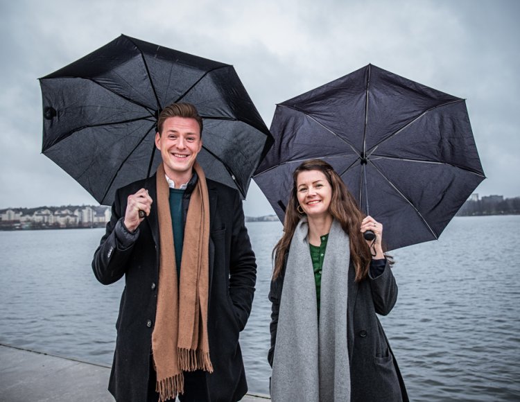 Ida Landin och Jonas Lundgren står utomhus under varsitt paraply med vatten i bakgrunden.