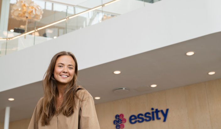 Emma Ottosson, som är Consumer Marketing Assistant på Essity står i företagets lobby. Hon ler mot kameran, och i bakgrunden syns Essitys logotyp på en vägg. 