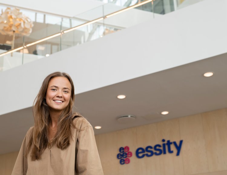 Emma Ottosson, som är Consumer Marketing Assistant på Essity står i företagets lobby. Hon ler mot kameran, och i bakgrunden syns Essitys logotyp på en vägg. 
