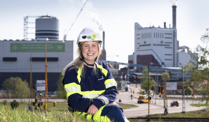 Gruppchefen Tina Jonasson sitter i gräset på en kulle framför BillerudKorsnäs stora fabriker.