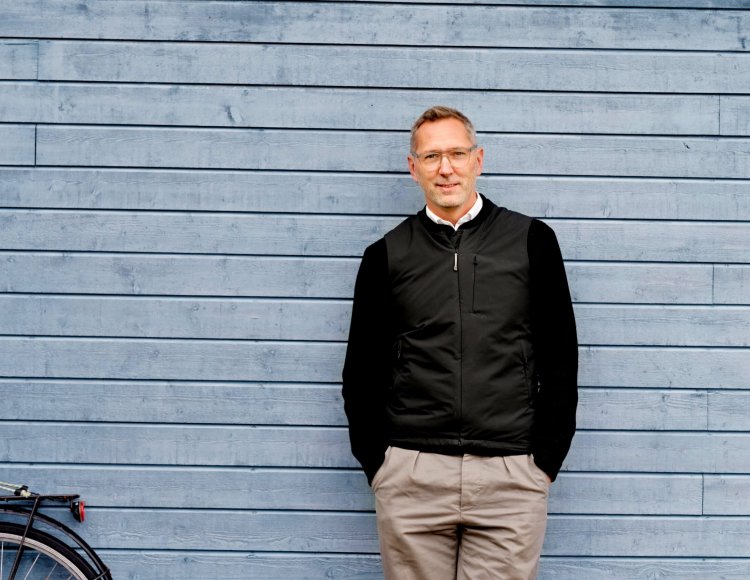 Anders på Ericsson står utomhus och lutar sig mot en blå träfasad. Han tittar in i kameran och ler. 