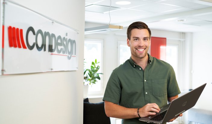 Tobias Elgström står på ett kontor, framför Condesign-logon med en dator i handen.