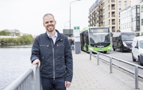 Fredrik Holmlund på Trafikverket står utomhus på en bro och håller i räcket. Han ler mot kamera. Bakom honom syns en grön buss från Skånetrafiken.

